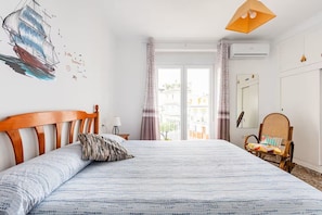 A bedroom with a bed, a wooden headboard, a rattan chair, a window with curtains, and a view of buildings outside. - Spacious and bright apartment with large private roof terrace-solarium. (Nerja)