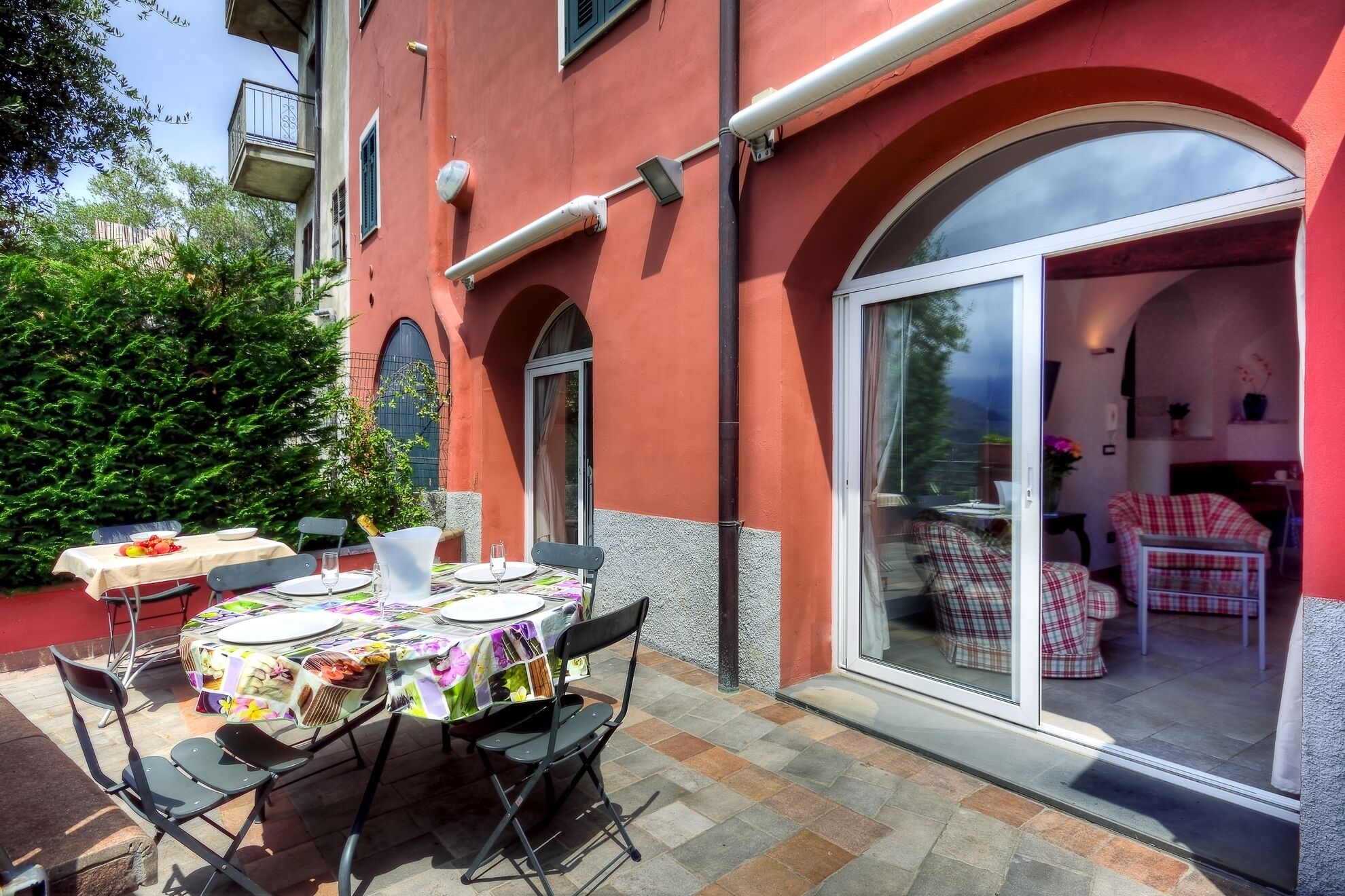 Un bâtiment rouge avec une table de patio dressée pour le dîner, une porte vitrée donnant sur un salon, et un arbuste vert en arrière-plan.