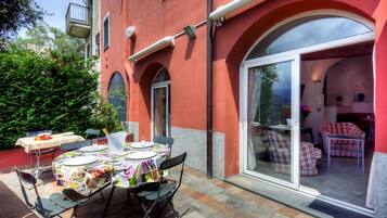 Un bâtiment rouge avec une table de patio dressée pour le dîner, une porte vitrée donnant sur un salon, et un arbuste vert en arrière-plan.