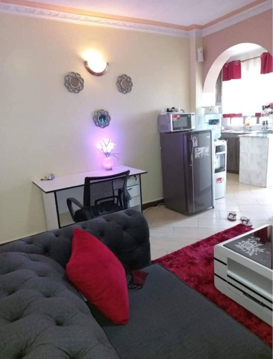 Un salon avec un canapé gris, un coussin rouge, une table à manger avec une chaise, un coin cuisine équipé d’appareils électroménagers et un petit îlot.
