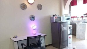 Un salon avec un canapé gris, un coussin rouge, une table à manger avec une chaise, un coin cuisine équipé d’appareils électroménagers et un petit îlot.
