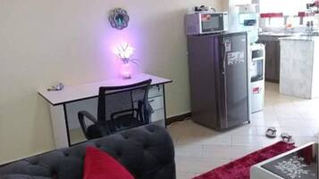 Un salon avec un canapé gris, un coussin rouge, une table à manger avec une chaise, un coin cuisine équipé d’appareils électroménagers et un petit îlot.
