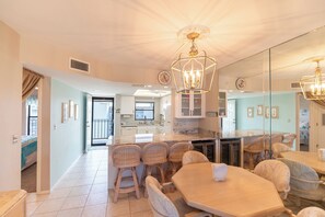 A spacious dining area with a large wooden table and wicker chairs, a kitchen with white cabinets, and a mirrored wall reflecting the room. - Fun In Sun On A Budget! 2B/2B Condo w/ Pool & Tennis! Walk to Beach! EC163 (Fort Myers Beach)