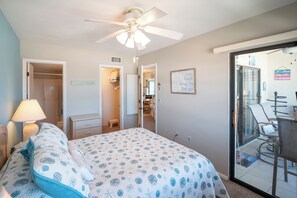 Ein Schlafzimmer mit einem Bett, einem Deckenventilator, einem Nachttisch, einer Lampe, einem Kleiderschrank und einer Glastür zum Balkon.