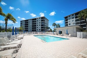 A swimming pool with lounge chairs and umbrellas, surrounded by a white fence, with buildings in the background. - Fun In Sun On A Budget! 2B/2B Condo w/ Pool & Tennis! Walk to Beach! EC163 (Fort Myers Beach)