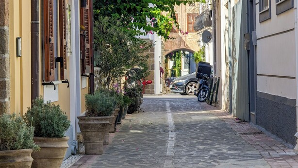 Una calle empedrada estrecha bordeada de macetas con plantas y coches aparcados.