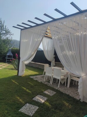 A covered outdoor seating area with white curtains, a grill, and a garden path. - Fethiye Göcek Villa Begonvilla (Fethiye)