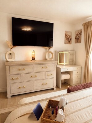 Un dormitorio con una televisión de pantalla grande montada en la pared, un tocador con espejo, un escritorio y una cama con cabecera.