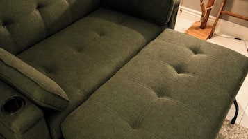 Un sofá verde con reposapiés y una mesita, ubicado cerca de una ventana con una lámpara.