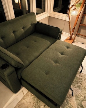 Un sofá verde con reposapiés y una mesita, ubicado cerca de una ventana con una lámpara.