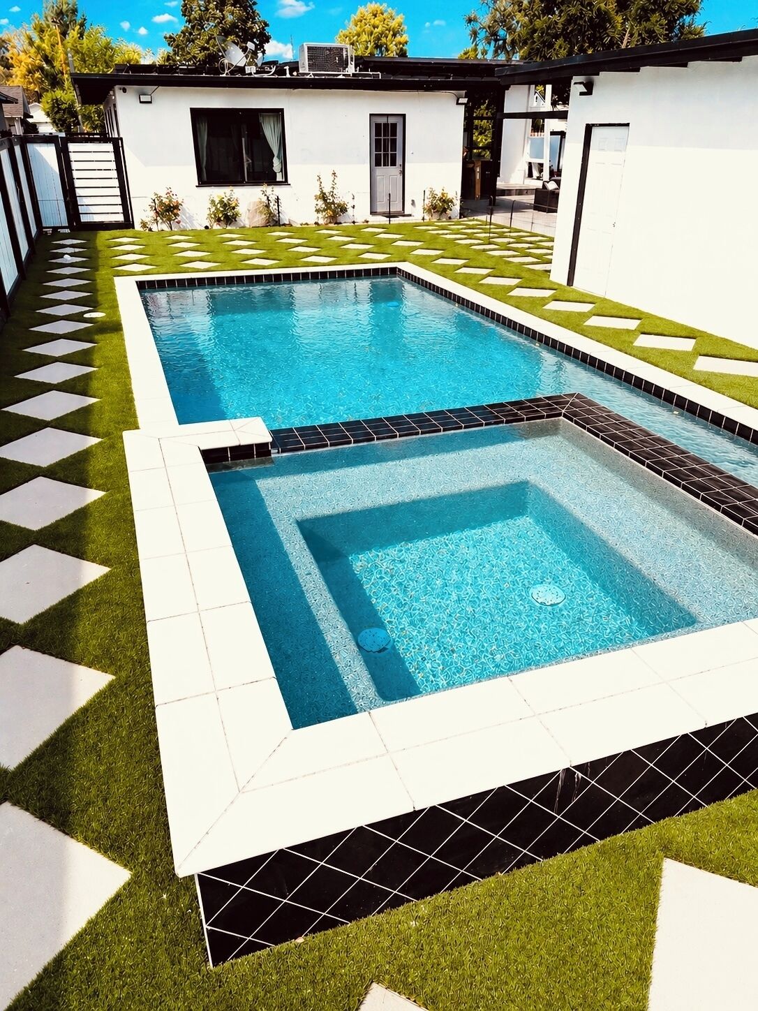 A swimming pool with a geometric tile pattern, surrounded by artificial grass and a white wall.