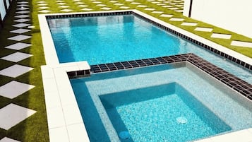 En swimmingpool med et geometrisk flisemønster, omgivet af kunstgræs og en hvid væg.