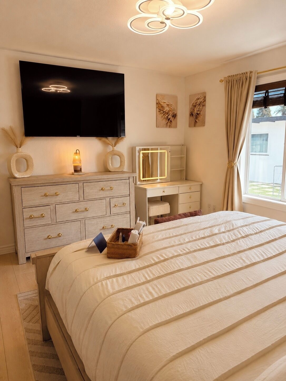 A bedroom with a bed, a dresser with drawers, a vanity with a mirror, a television mounted on the wall, and a window with curtains.