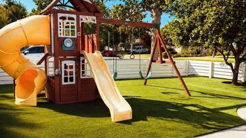 Un parque infantil con tobogán y columpios, rodeado de césped cuidado y árboles.