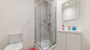 Un baño con cabina de ducha de vidrio, inodoro, alfombra roja, lavabo blanco con espejo y un armario.