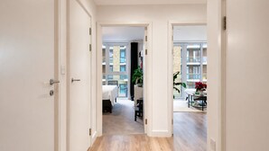 A hallway leading to a bedroom with a bed, a desk, and a chair, and a dining area with a table and chairs. - Modern Riverside Apartment | Balcony | Elizabeth Line (London)