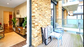 A room with a brick wall, a balcony with chairs, a wooden floor, and a large window. - Modern Riverside Apartment | Balcony | Elizabeth Line (London)