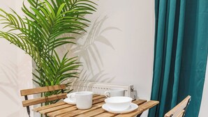Ein Holztisch mit weißen Tassen und einer Schale, ein Stuhl, eine große grüne Pflanze und blaue Vorhänge.