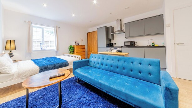 A modern living room with a blue sofa, a small coffee table, and a kitchen area with cabinets and appliances. - Cosy Serene Flat near the City Centre (Kent)