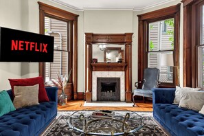 Ein Wohnzimmer mit Kamin, zwei blaue Sofas, einem Couchtisch und einem Fernseher, der 'NETFLIX' anzeigt.