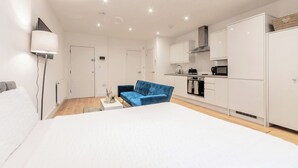 Una habitación de hotel moderna con una cama grande, un sofá azul, una kitchenette y un techo blanco con iluminación empotrada.