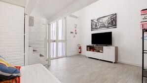 A room with white brick walls, a white ceiling, a flat-screen TV on a wooden TV stand, a fire extinguisher, and a staircase leading to an upper level. - Holiday Home 'Ciccina' with Wi-Fi and Air Conditioning (Palermo)