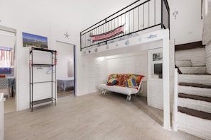 Ein Loft-Zimmer mit weißer Backsteinwand, einem weißen Sofa mit bunten Kissen, einem Metallregal und einer Treppe mit schwarzer Geländer.