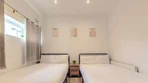 Een kamer met twee enkele bedden, een klein houten nachtkastje met een plantje en een raam met gordijnen.