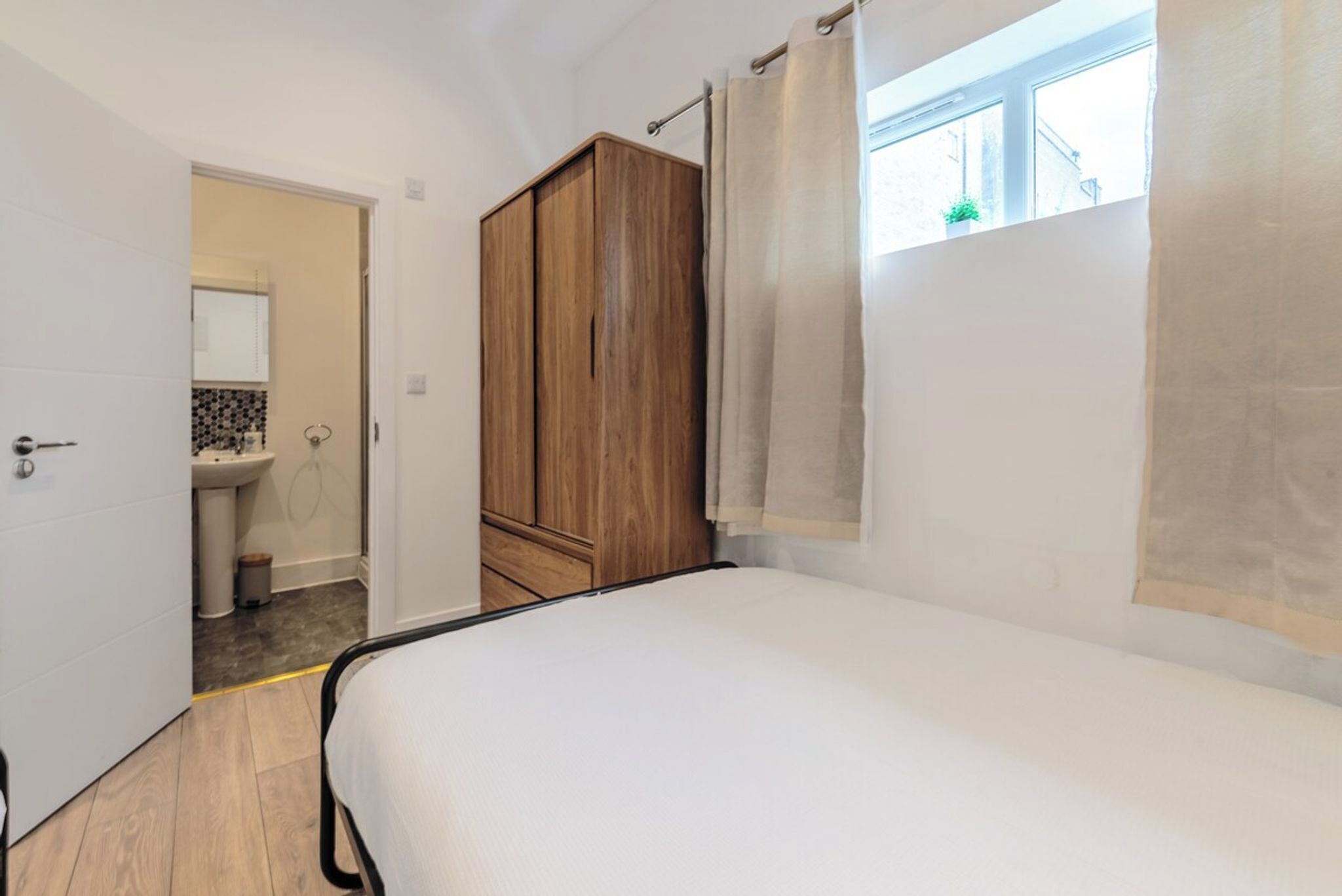 A bedroom with a bed, a wardrobe, a window with curtains, and a bathroom visible in the background.