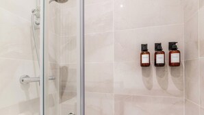 Et bruseområde med glasvægge, håndbruser og en glasdør. Tre shampoo-flasker er monteret på væggen.
