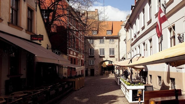 Une ruelle étroite pavée, bordée de bâtiments des deux côtés, avec des tables en terrasse et un drapeau.