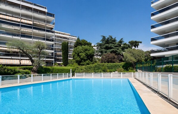 En swimmingpool med en hvid terrasse, omgivet af grøn bevoksning og moderne bygninger.