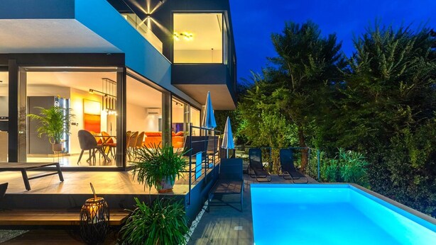 Una casa moderna de dos pisos con fachada de vidrio, mobiliario exterior y una piscina.