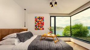 Une chambre moderne avec un grand lit, une vue sur la mer et un tableau abstrait aux couleurs vives.