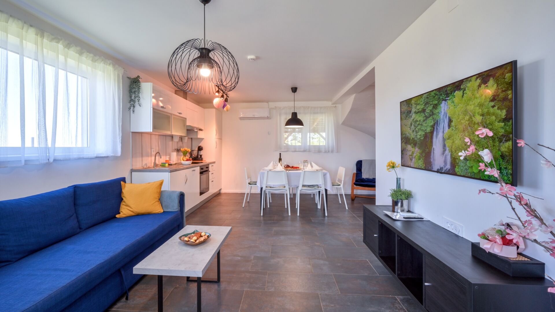 Un appartement moderne avec un canapé bleu, une table à manger dressée pour quatre personnes, une cuisine en arrière-plan et un grand tableau représentant un paysage naturel accroché au mur.
