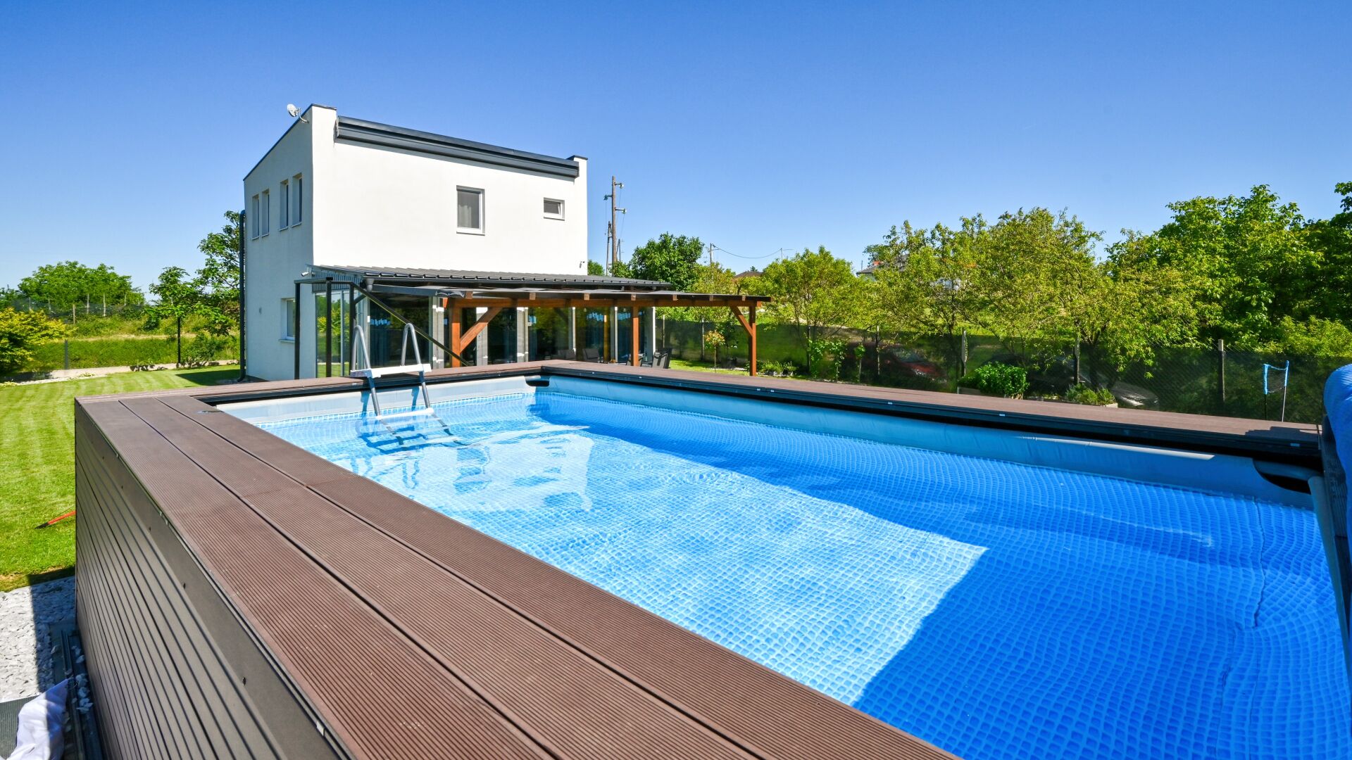 Une piscine avec une terrasse en bois, un bâtiment blanc et un espace couvert avec des chaises.
