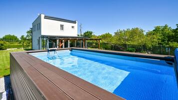 Une piscine avec une terrasse en bois, un bâtiment blanc et un espace couvert avec des chaises.