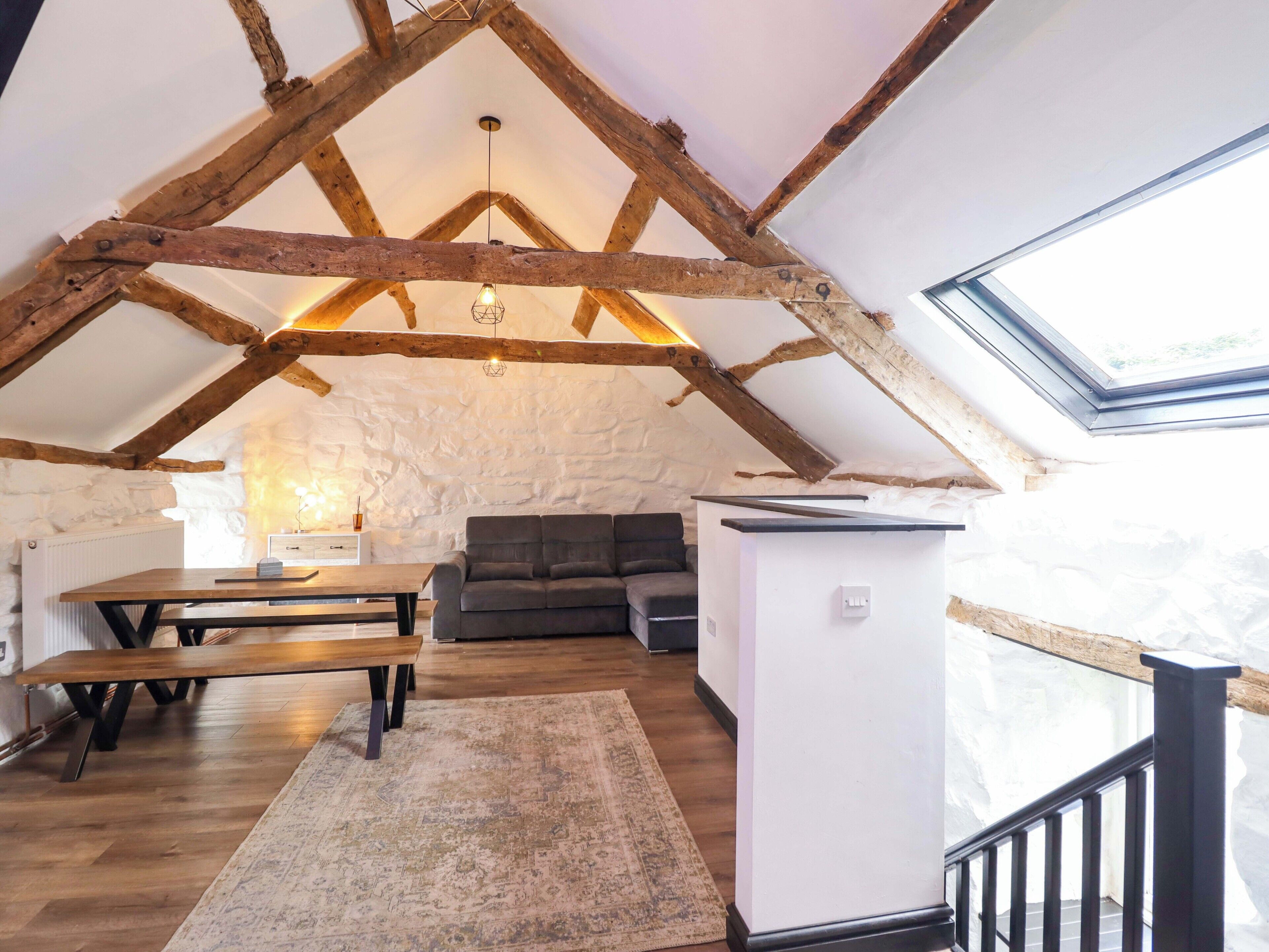 Un espacio tipo loft con vigas de madera a la vista, una pared de piedra, una mesa de madera con bancos, un sofá, un mueble blanco pequeño y un tragaluz.