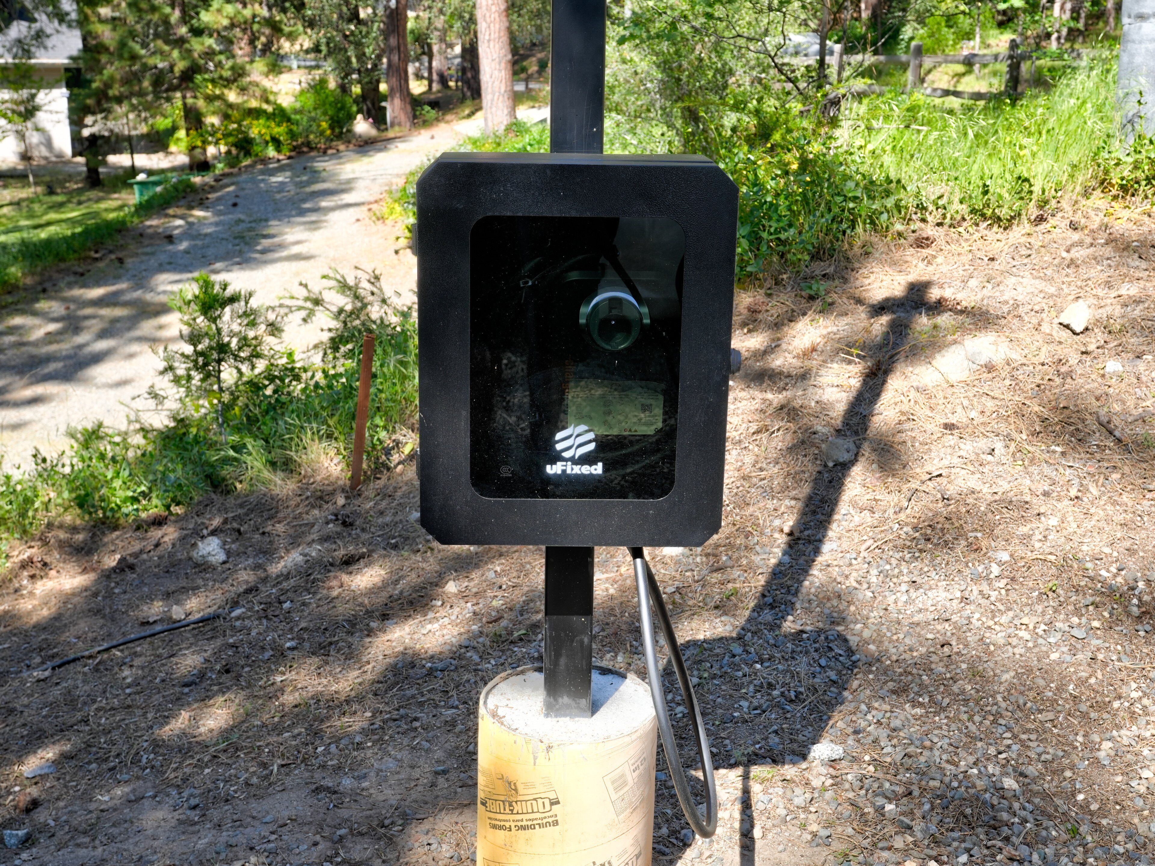 Une caméra de surveillance extérieure noire, fixée sur un poteau doté d’une base métallique, est entourée d’un environnement naturel composé d’arbres et d’un chemin de gravier.