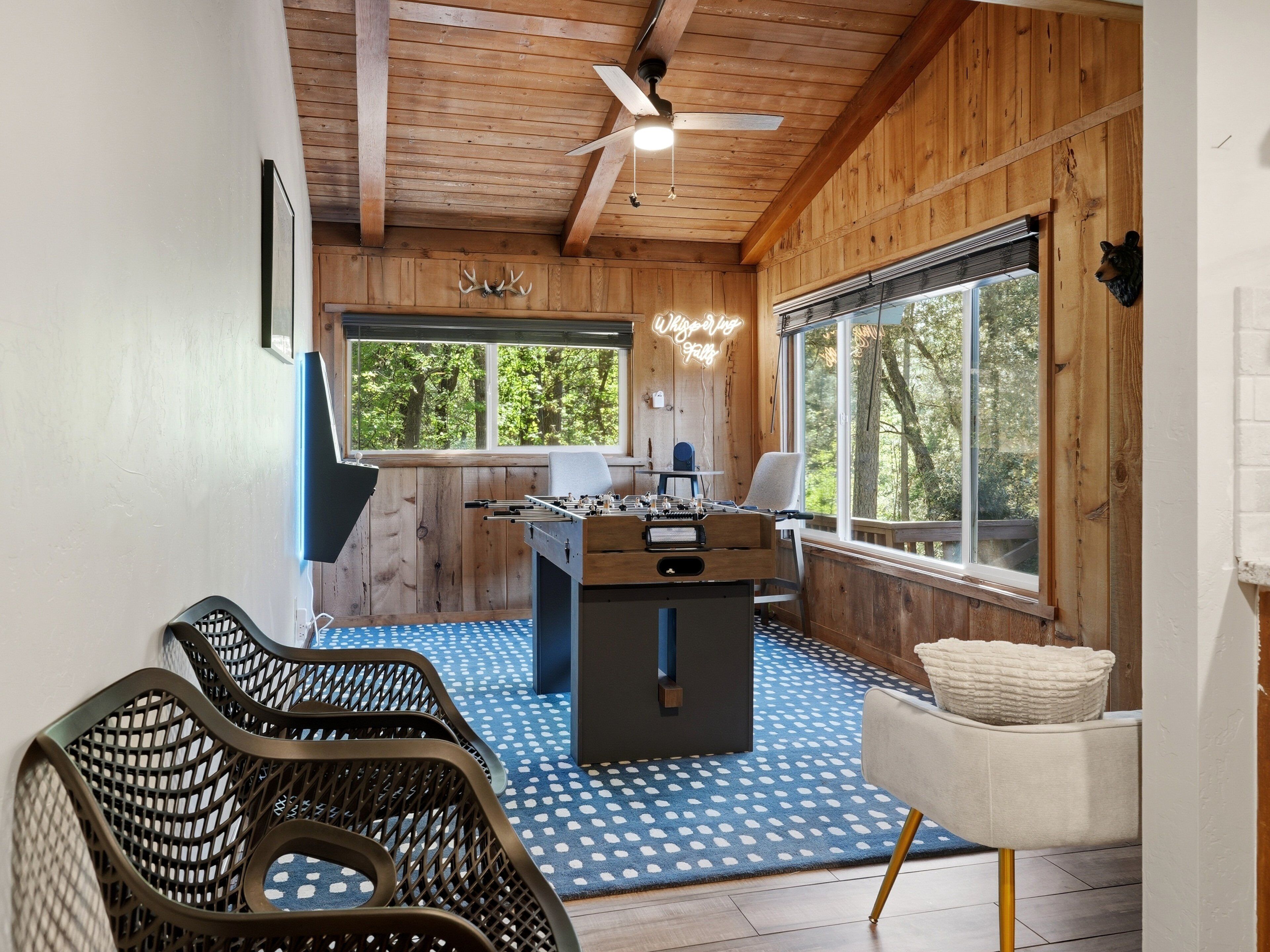 Una habitación acogedora con techo de madera, una mesa de fútbol de mesa, una ventana con vista a árboles y una alfombra azul estampada.