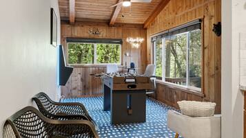 Una habitación acogedora con techo de madera, una mesa de fútbol de mesa, una ventana con vista a árboles y una alfombra azul estampada.
