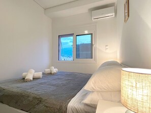 A bedroom with a bed, a window with a view, an air conditioner, and a lamp. - Bay View - Bright Modern Penthouse in Center (Kalamata)