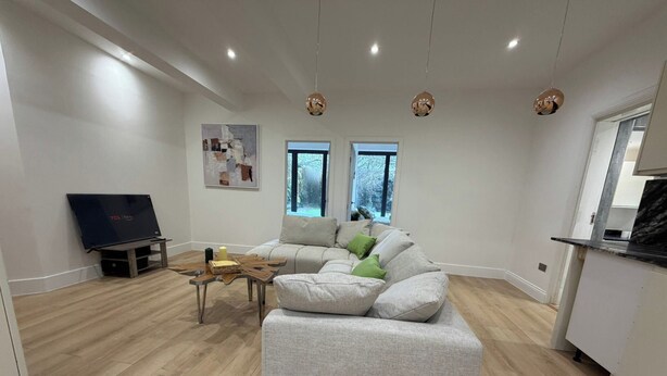 A modern living room with a grey sectional sofa, a wooden coffee table, a flat-screen TV, and a kitchen area with a marble countertop. - 2 Bed - Sleeps 5 - Garden - Hot-tub (London)