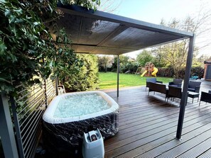 A hot tub on a wooden deck with a covered patio area, surrounded by greenery and a playhouse in the background. - 2 Bed - Sleeps 5 - Garden - Hot-tub (London)
