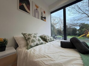 A bedroom with a large bed, two pillows, a nightstand with a plant, and a window with a view of a playground. - 2 Bed - Sleeps 5 - Garden - Hot-tub (London)