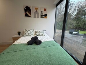 A bedroom with a large window, a bed with green bedding, a nightstand with a plant, and abstract wall art. - 2 Bed - Sleeps 5 - Garden - Hot-tub (London)