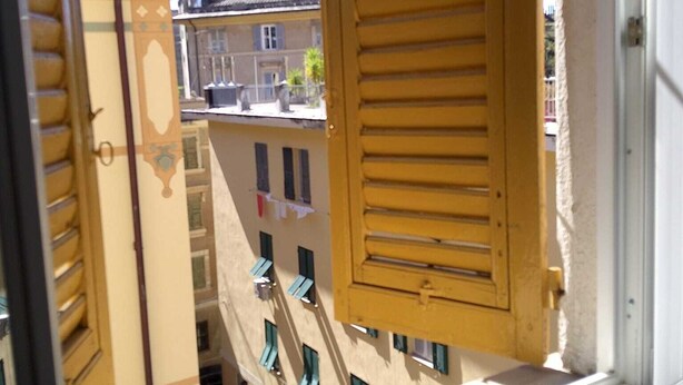 Una ventana con un persiano de madera entreabierto, mostrando una vista de un edificio con balcones y ropa tendida.