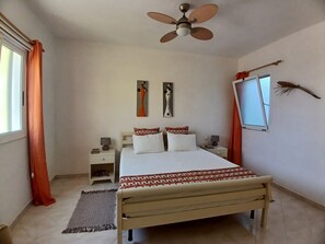 Ein Schlafzimmer mit einem Bett, einem Deckenventilator, Kunstwerken an der Wand, einem Fenster und einem Nachttisch mit einer Lampe.