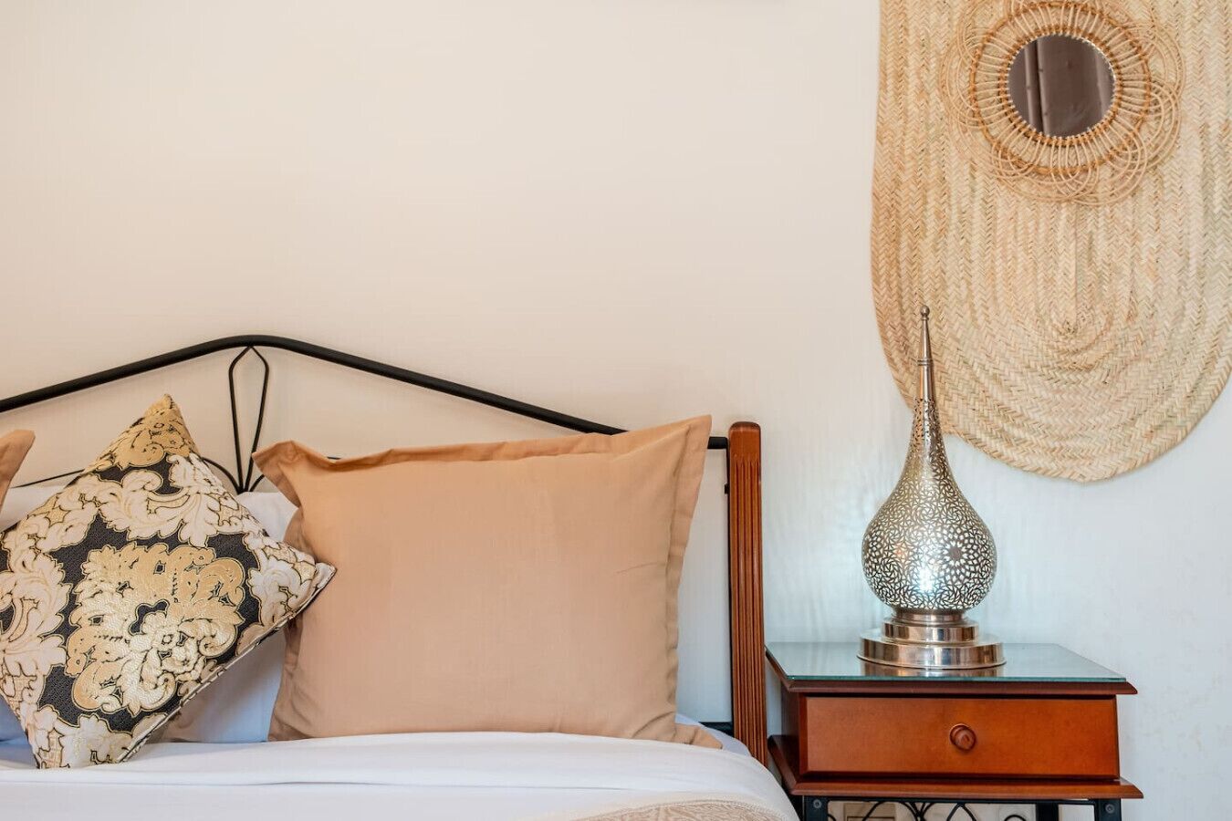 A bed with a headboard, two pillows, a bedside table with a lamp, and a woven wall hanging.
