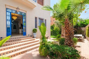 A building with a blue door, steps leading to the entrance, a palm tree, and a garden with various plants. - Jolie villa 4 chambres à 10 minutes centre (Marrakesh)